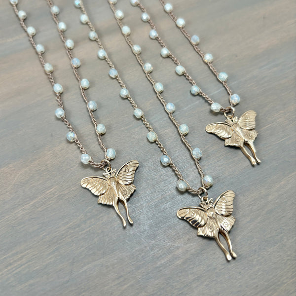 Three gold fairy luna moth necklaces with  small white pearl-like beads on a textured gray wooden surface.