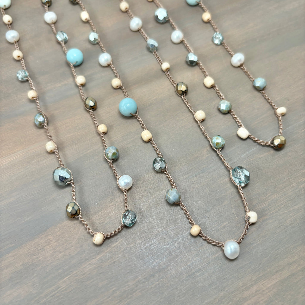 A close-up of a beaded necklace laid flat on a wooden surface. The necklace features a copper-toned chain with alternating light blue turquoise beads, white pearl-like beads, faceted gray-green beads, and small metallic gold and bronze beads.
