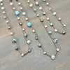 A close-up of a beaded necklace laid flat on a wooden surface. The necklace features a copper-toned chain with alternating light blue turquoise beads, white pearl-like beads, faceted gray-green beads, and small metallic gold and bronze beads.