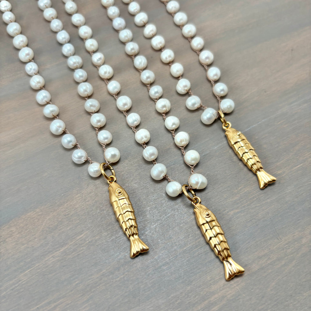 Three gold fish-shaped pendant necklaces with white pearl-like beads, arranged diagonally on a dark wood surface.