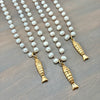 Three gold fish-shaped pendant necklaces with white pearl-like beads, arranged diagonally on a dark wood surface.