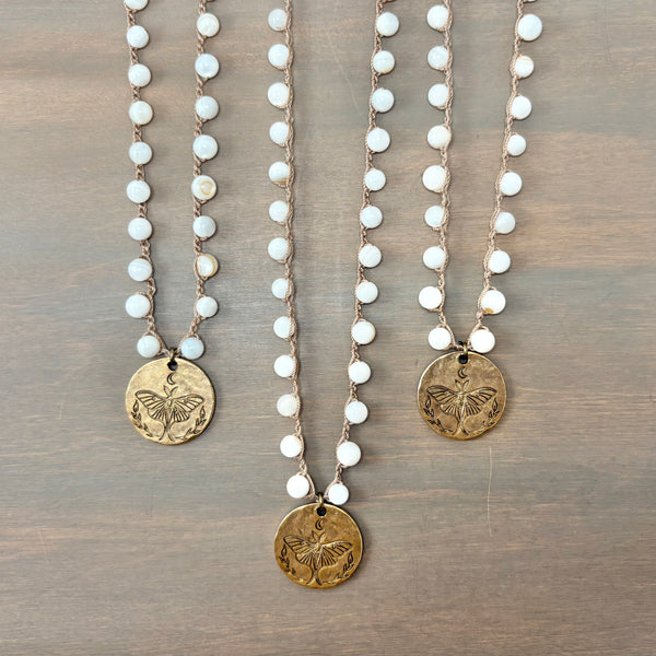 Three bohemian-style beaded necklaces with white round beads and antique bronze butterfly coin pendants, arranged on a brown wooden surface.